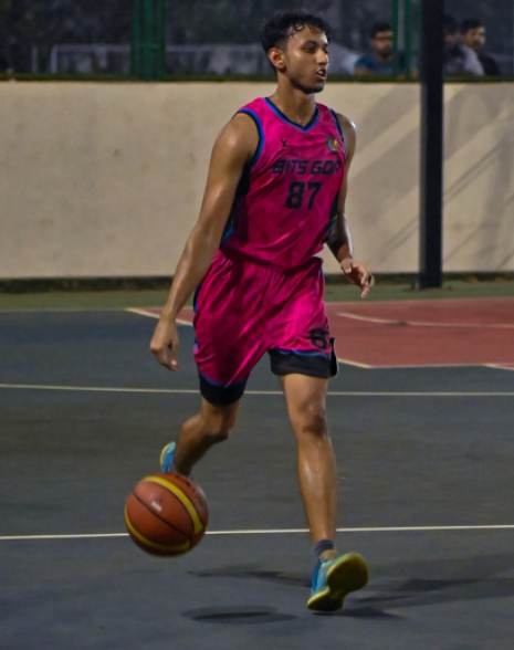 Yash Lothe playing basketball in a pink BITS Goa jersey, dribbling up the court under lights.
