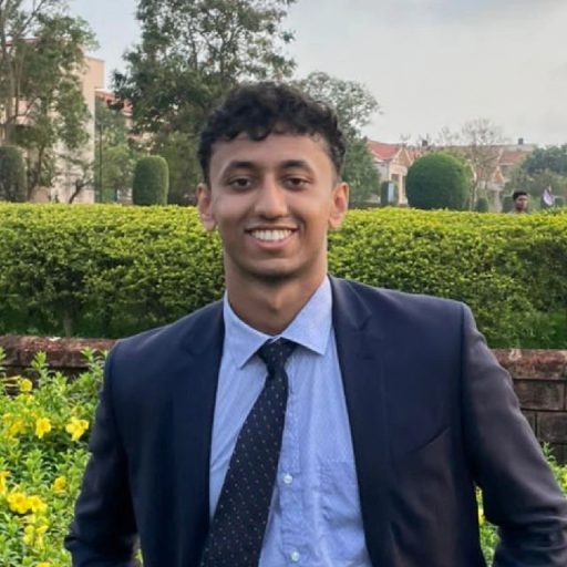 Yash Lothe, smiling, wearing a navy suit and light blue shirt, photographed outdoors in front of a hedge.